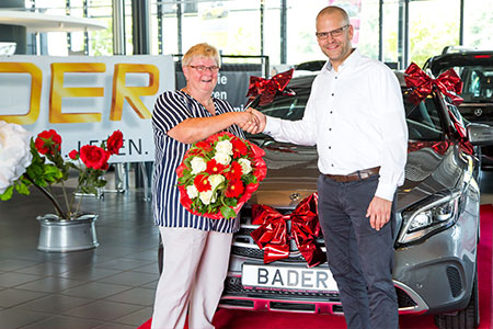 Gewinnspielübergabe vor einem mit Schleife geschmückten Mercedes-Benz mit BADER-Kennzeichen; zwei Personen geben sich die Hand, eine hält einen großen Blumenstrauß.