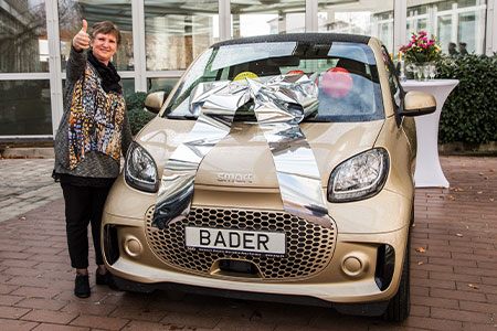 Gewinnspielübergabe vor einem mit großer silberner Schleife geschmückten Smart mit BADER-Kennzeichen; eine Frau steht daneben und zeigt jubelnd den Daumen nach oben.